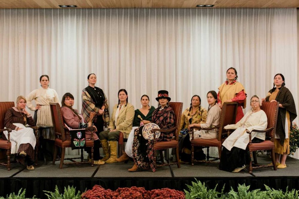 Indigenous women dressed in traditional clothing sitting on a stage in chairs