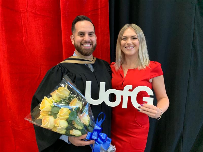 A graduate holds flowers alongside a woman holding a UofG sign