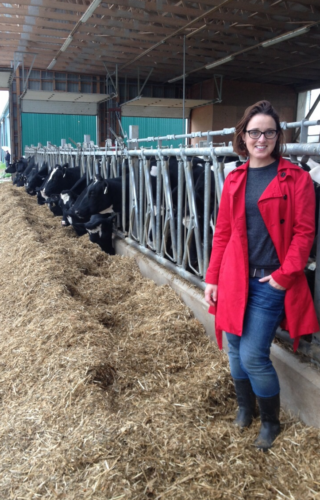 Prof. Christine Baes leaning against a railing with dairy cattle feeding on hay in the background