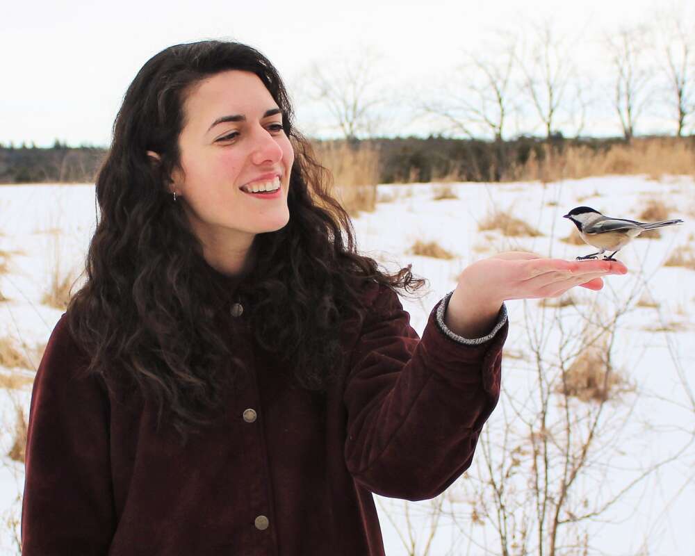 Hayley Wilson stands in a field feeding a chickadee from her hand.