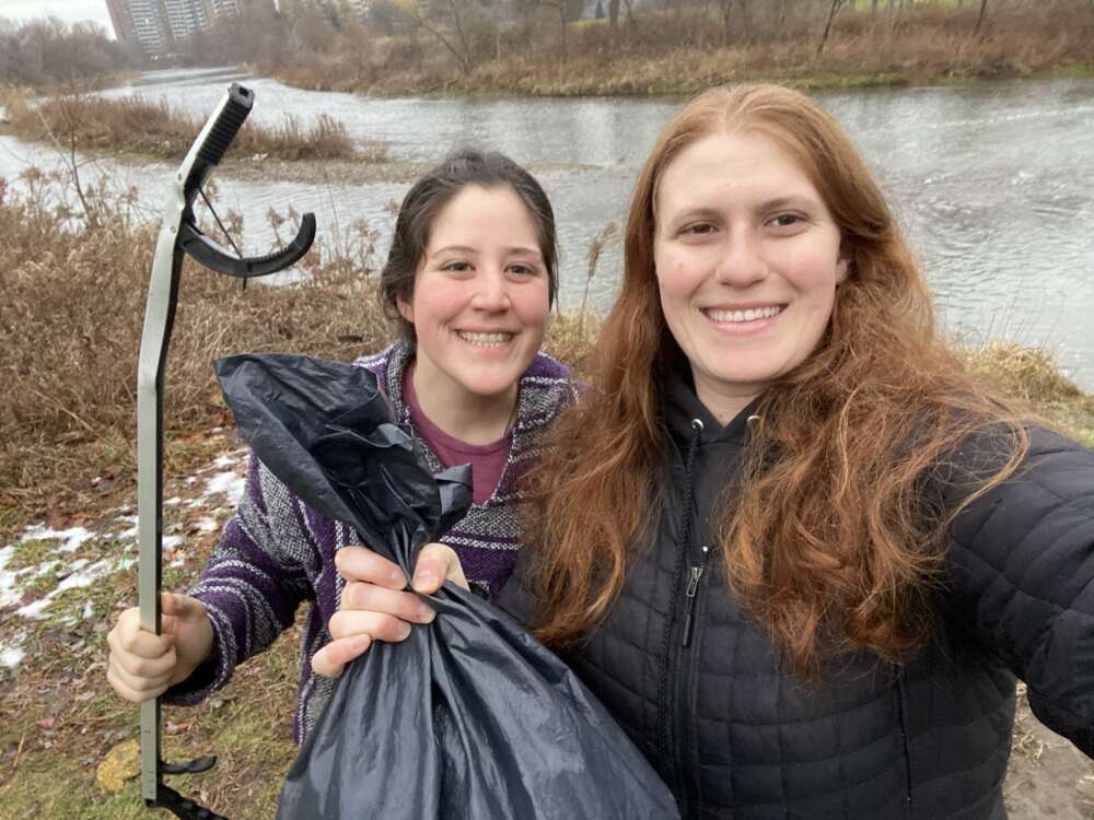 Two women stand outdoors, one on left holding a grabber, the one on right holding a garbage bag