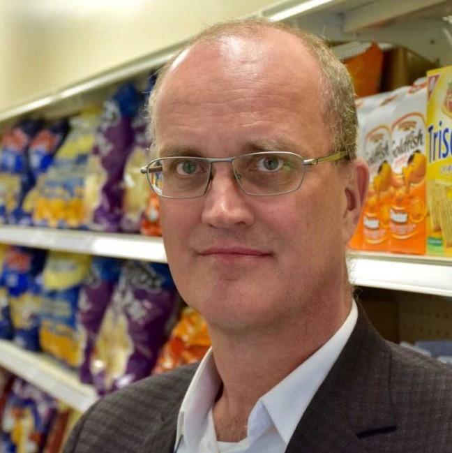 Dr. Mike von Massow poses for a headshot in front of a shelf of potato chips and goldfish at a grocery store.