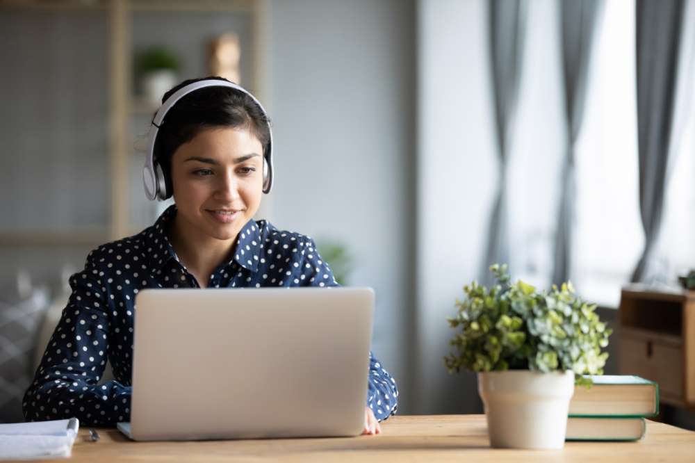 A woman wearing headphones smiles as she looks at a laptop at a desk