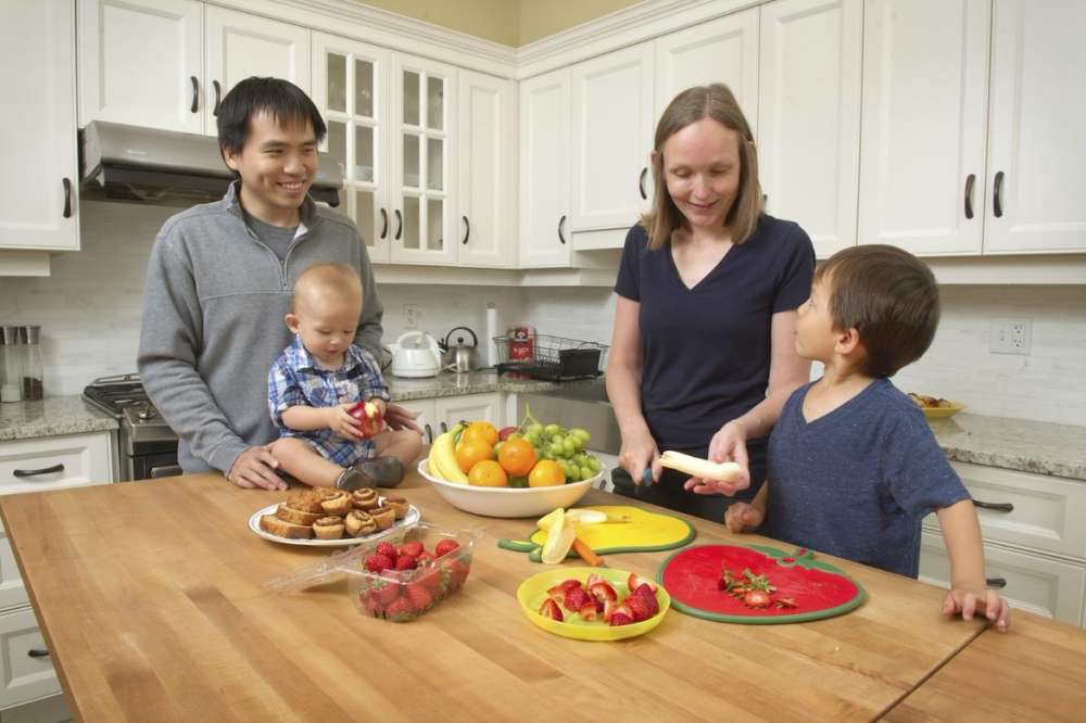 family with two young children around a kitchen counter with vegetables and fruit cut up on plates