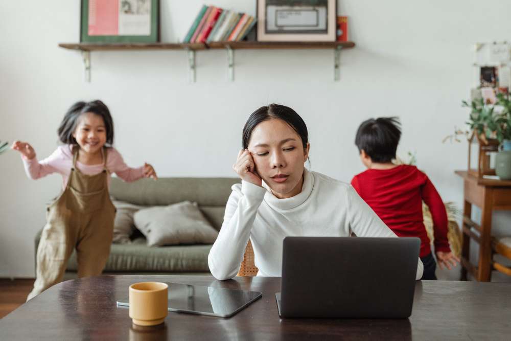 A tired-looking mother sits at a laptop while two children run in circles behind her.