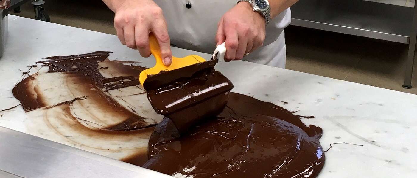 A chef uses a spatula to shear melted chocolate on a marble slab