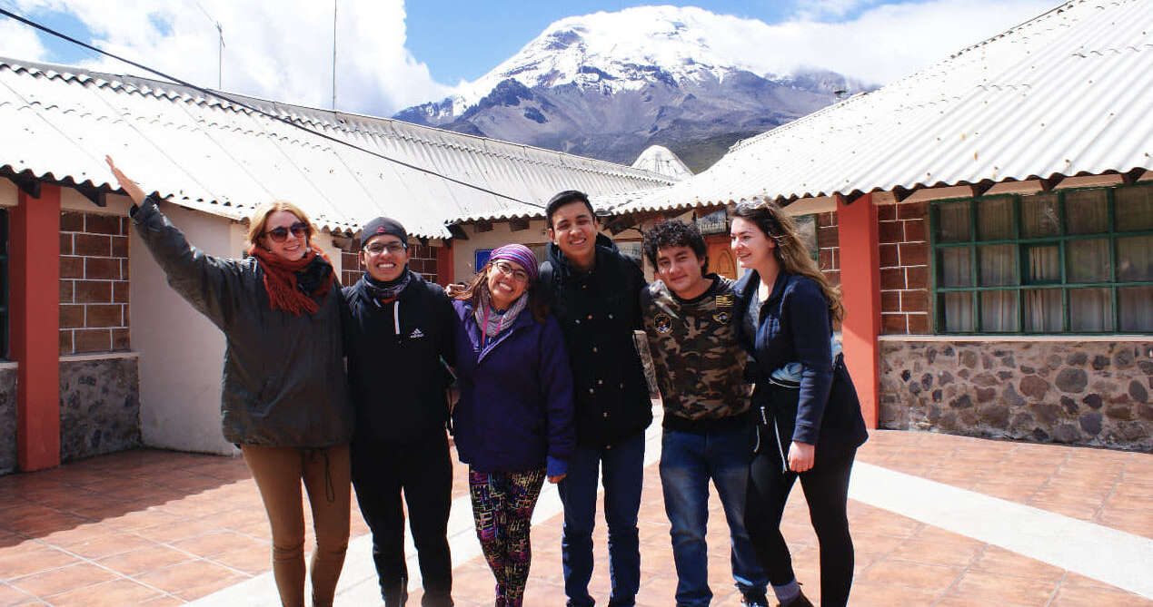Six people wearing warm clothes stand in a courtyard in front of a snow-covered mountain