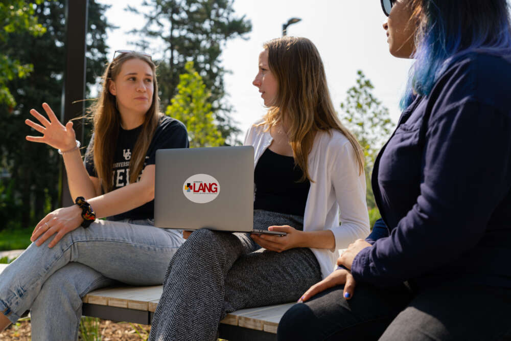 three female students sitting on a concrete bench outdoors interacting with one another. One student has a lap top sitting on her knee and the Lang logo is on the top of the laptop