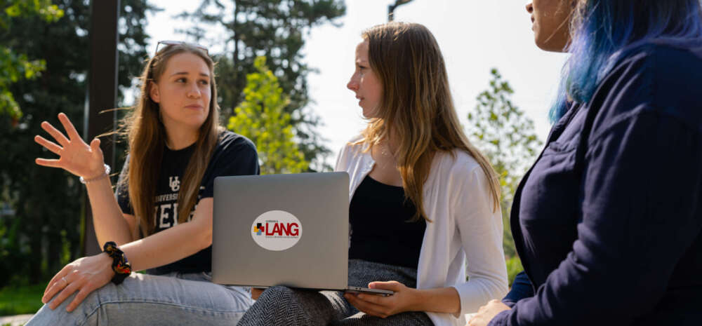 three female students sitting on a concrete bench outdoors interacting with one another. One student has a lap top sitting on her knee and the Lang logo is on the top of the laptop