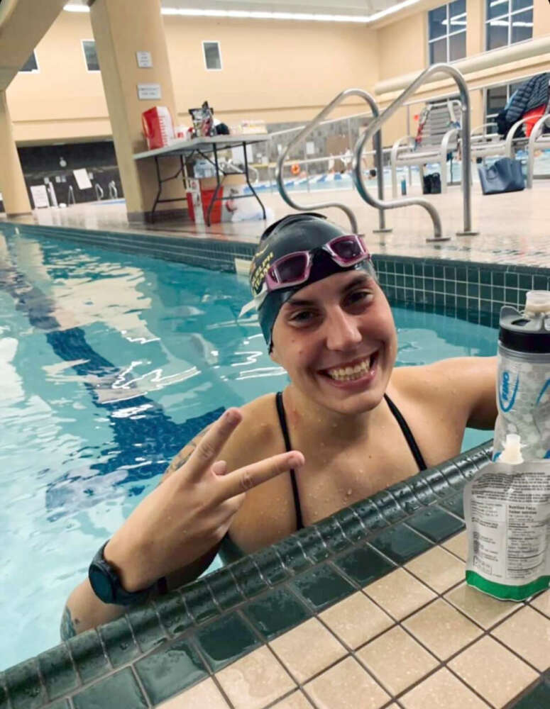 A woman in a swimcap stands in a pool and smiles for the camera from