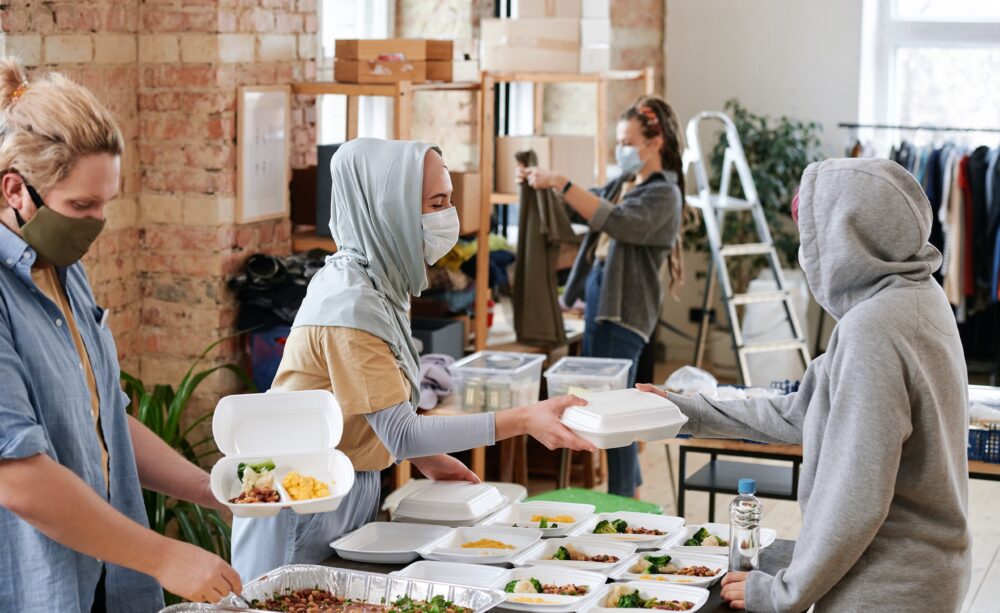 Two people wearing masks fill takeout containers and pass one to a person wearing a hoodie