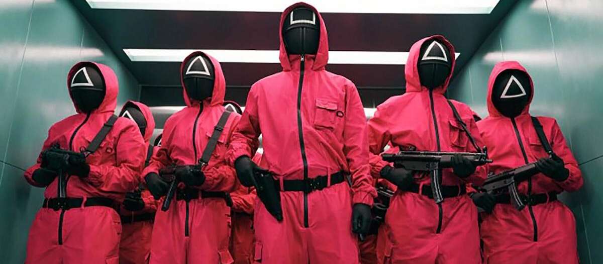 Five Squid Game workers in red jumpsuits and black masks stand in a line holding guns