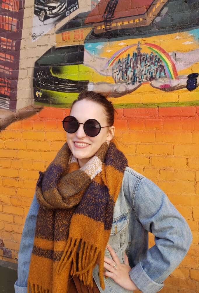 Emma Hak-Kovacs wears sunglasses and stands before a brick wall of graffiti