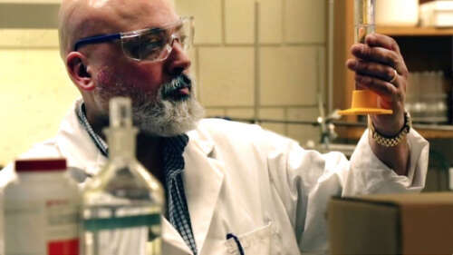 Man with beard and wearing safety glasses and lab coat stands amid equipment in a lab