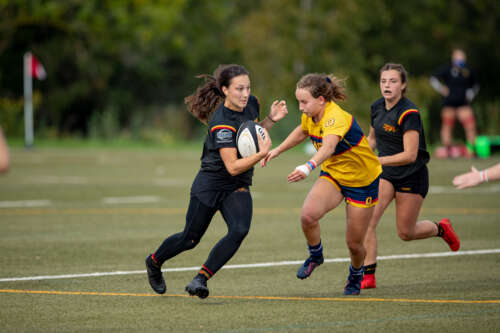 University of Guelph's Talia Hoffman dodges a tackle, while holding the rugby ball, from a Queen's University rugby player, in a yellow jersey, during a game.
