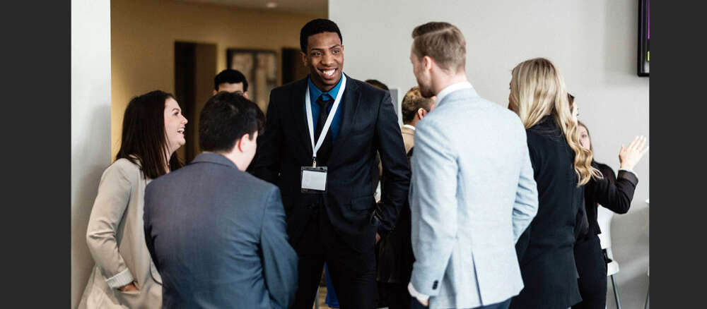 Several young people wearing suits stand in a circle laughing at a pre-pandemic gathering