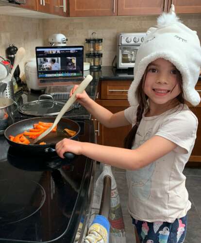 a girl wearing a unicorn cat smiles at the camera while stirring carrots in a pan