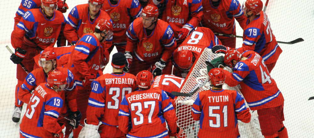 Russian Olympic Hockey players, wearing vibrant red jerseys, huddle around the net