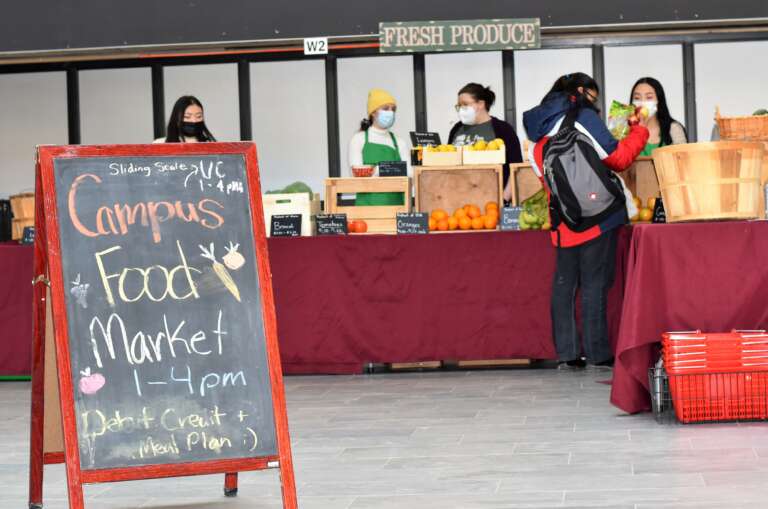 Siswa berdiri di meja dengan kotak produk.  Papan lipat di depan bertuliskan: Campus Food Market 13:00-16:00