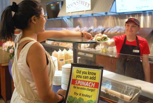 A woman at a sandwich bar takes a wrap from an employee