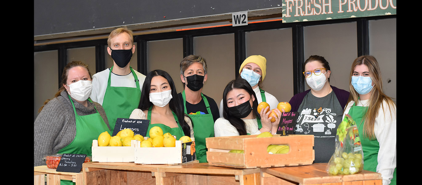 Eight people wearing masks and green aprons stand behind crates of produce