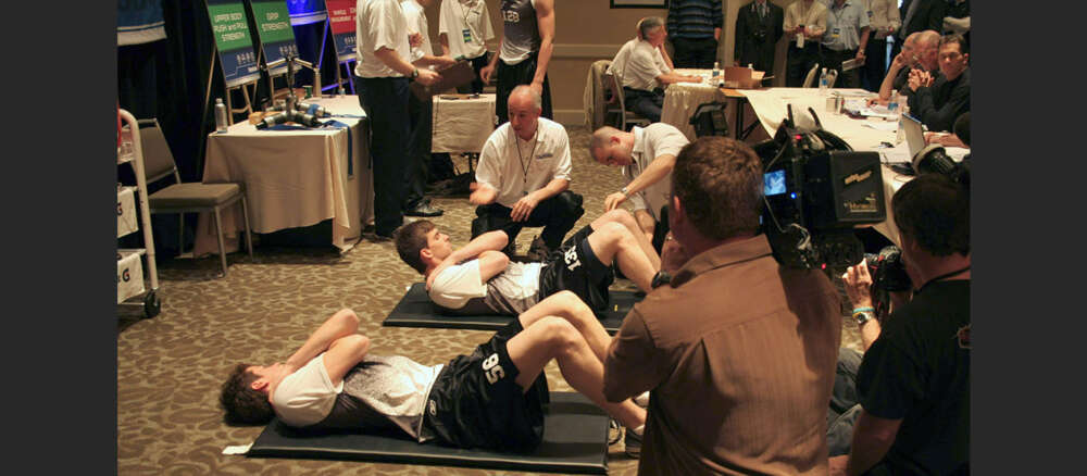 A coach speaks to 3 hockey players lying on mats doing situps while a videographer takes video