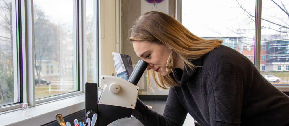 Danielle Nowosad in black sweater leans to peer into a microscope in front of a window.