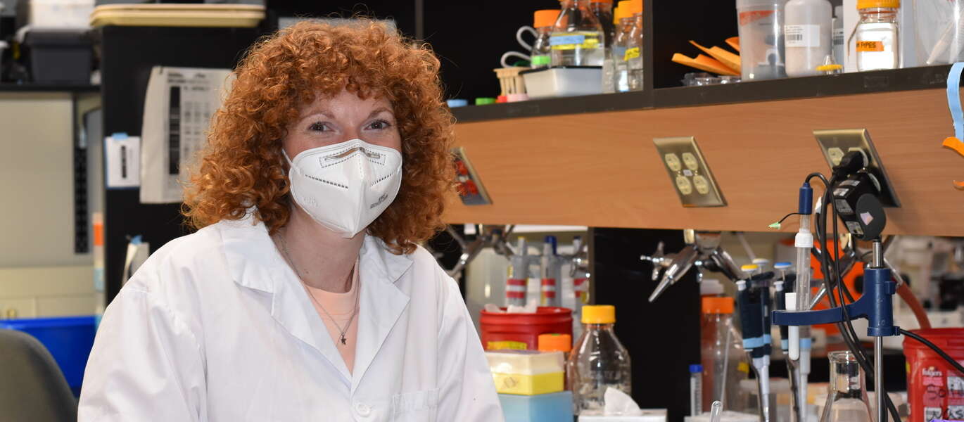 Dr. Jennifer Geddes-McAlister in lab coat sits at station in science lab.