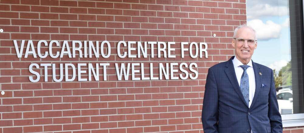 Former U of G president Franco Vaccarino stands against a brick wall with a sign that reads 'Vaccarino Centre for Student Wellness'