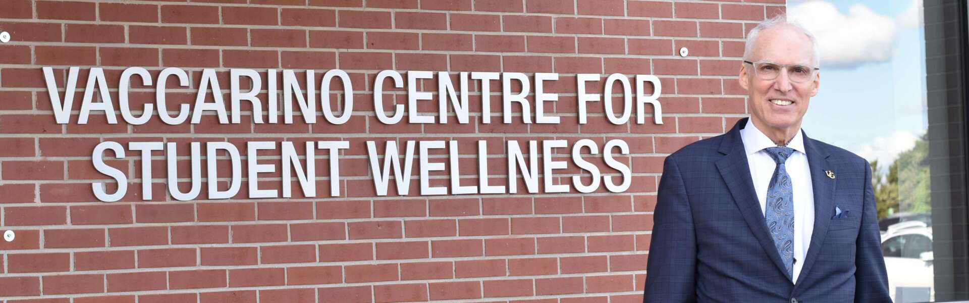 Former U of G president Franco Vaccarino stands against a brick wall with a sign that reads 'Vaccarino Centre for Student Wellness'
