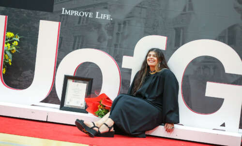 A graduate in conovcation robes sits beside their degree and a bouquet of flowers on a large U of G sign