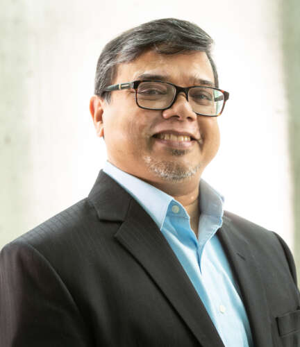 Dr. Sourav Ray stands in blue collared shirt and suit jacket against white background.