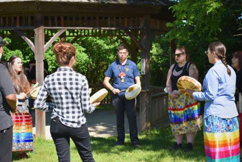 A group of people stand in a circle holding and playing hand drums on the grounds of The Arboretum.
