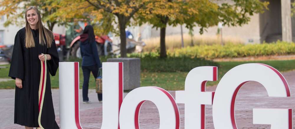 A graduate wearing convocation robes stands next to a giant UofG sign