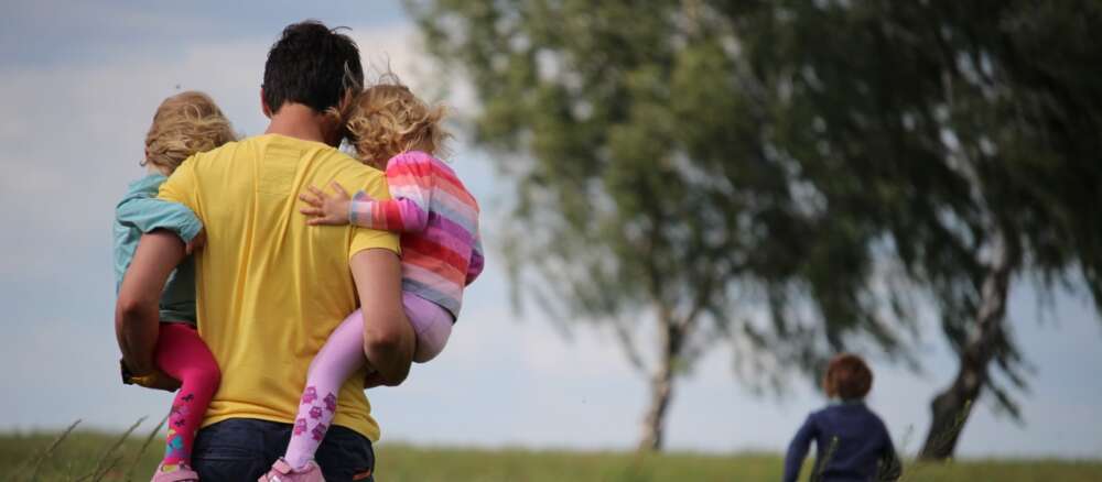 man carrying 2 young children on field of red flowers