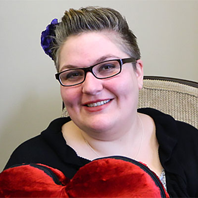Dr. Ruth Neustifter poses for a headshot against a grey wall, and is holding a red plush heart.