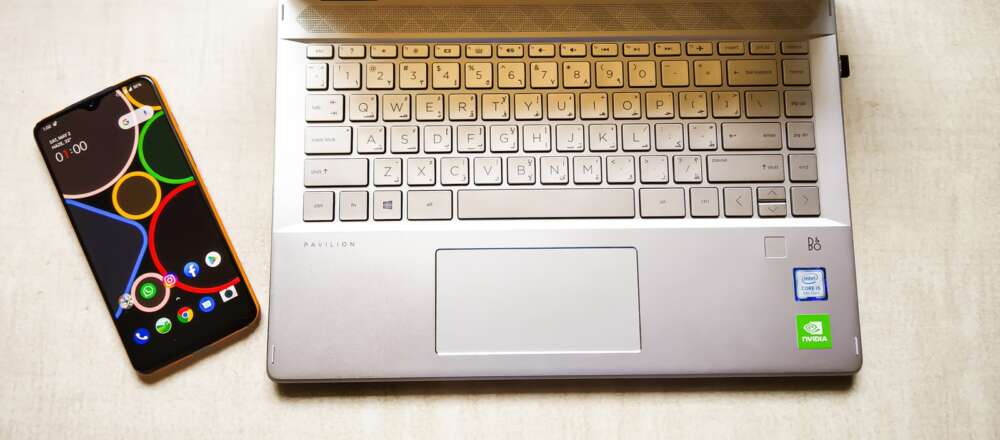 A phone set to its homescreen sits next to an open laptop on a table.