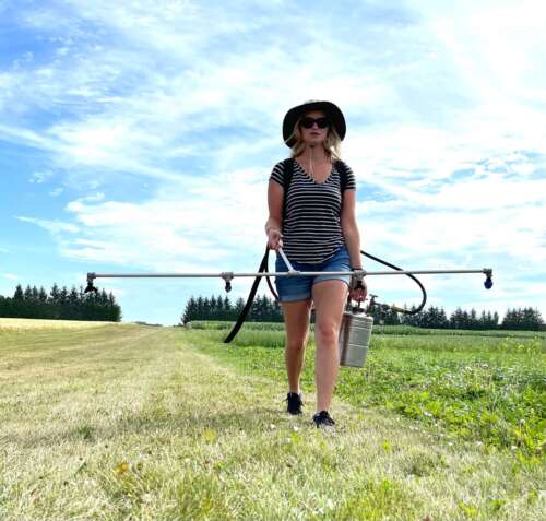 A woman earing a large brimmed hat walks along a field with a 4-nozzle sprayer and a canister