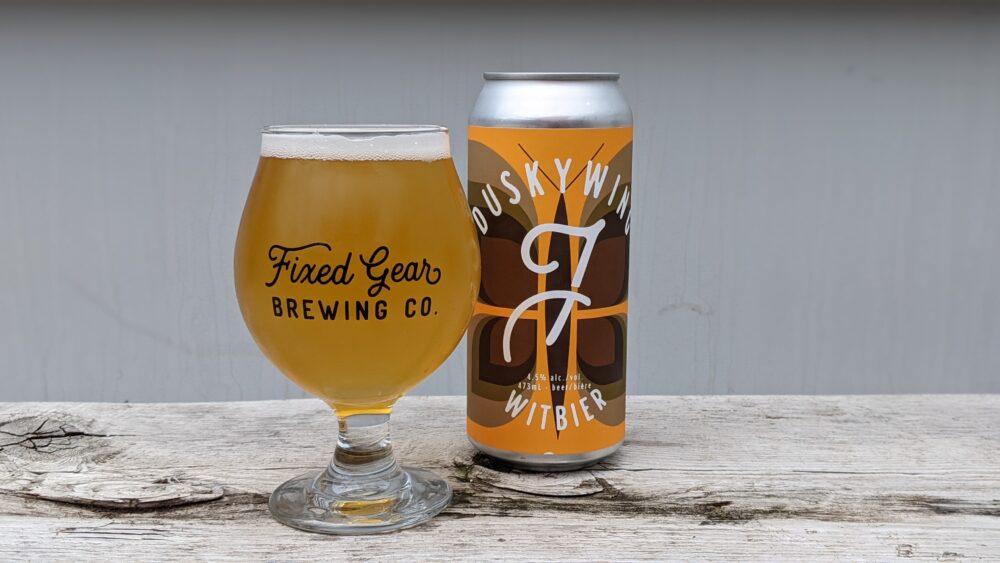 A glass of a yellow beer sits on a wooden table next to a silver can with a brown butterfly on an orange label.