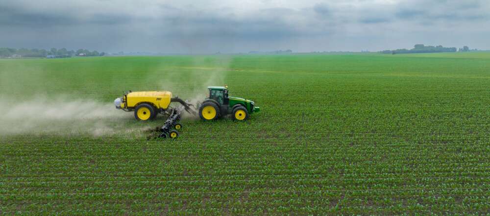 A green tractor with a yellow tank in a green field. The tractor is on the left side of the image and is moving towards the right side. Behind it is a plume of a white substance.