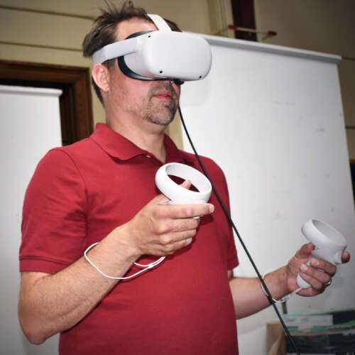 Dr. Peter Kuling in red collared t-shirt stands in VR headset and hand controls in front of open door at U of G