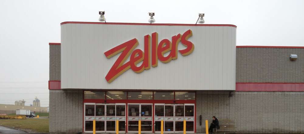 An entrance to a Zellers in a grey brick building. Above the doors is a white panel with the a large red sign proclaiming Zellers.