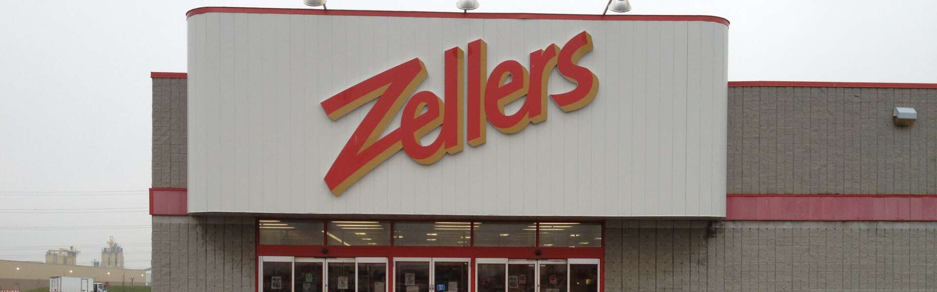 An entrance to a Zellers in a grey brick building. Above the doors is a white panel with the a large red sign proclaiming Zellers.