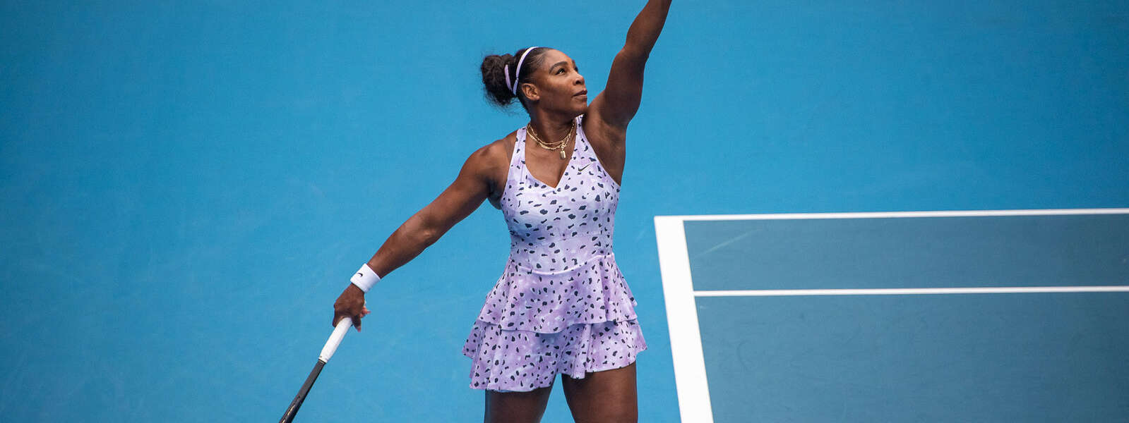 Serena Williams wearing a purple outfit serves on a blue tennis court.