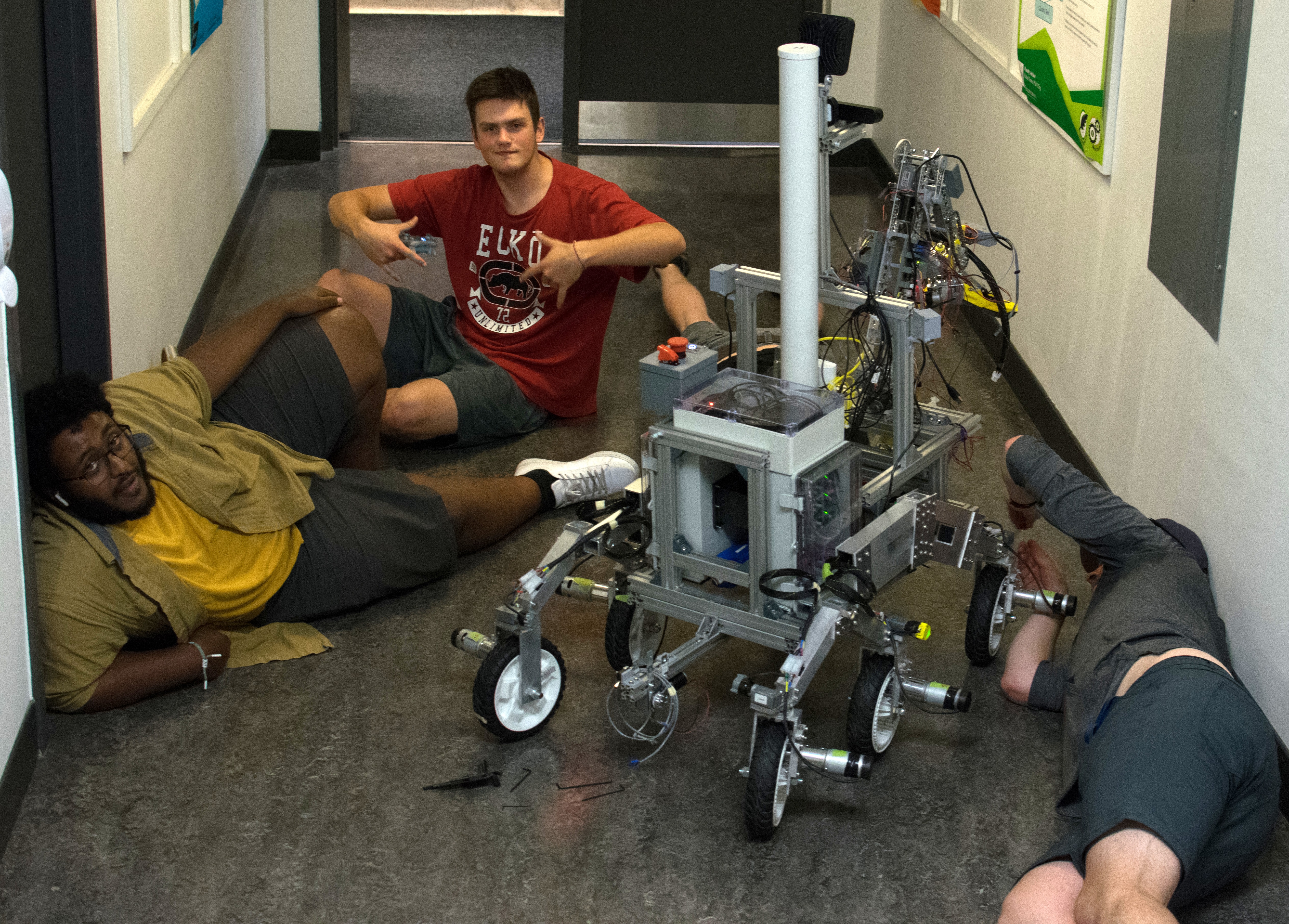 three people sit and lie on the floor of a university hallway and adjust a rover vehicle