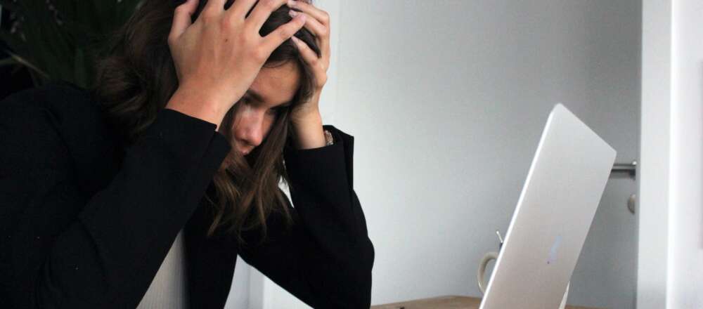 A person in black long sleeve shirt sits in front of their laptop. Their face is covered by their hands which are gripping their hair in a gesture expressing stress.