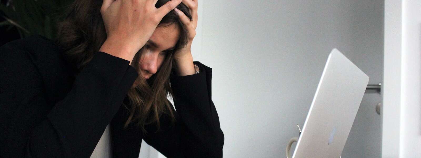 A person in black long sleeve shirt sits in front of their laptop. Their face is covered by their hands which are gripping their hair in a gesture expressing stress.