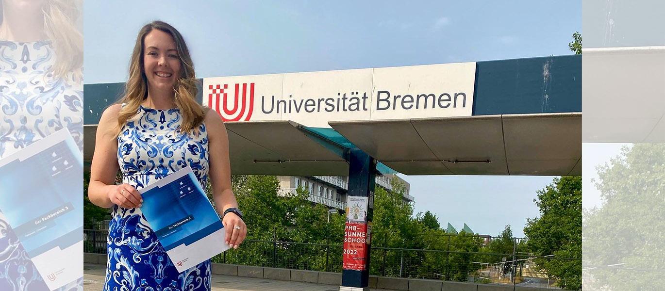 Dr. Becky Breau stands on campus at the University of Bremen in blue and white dress holding press materials for the school