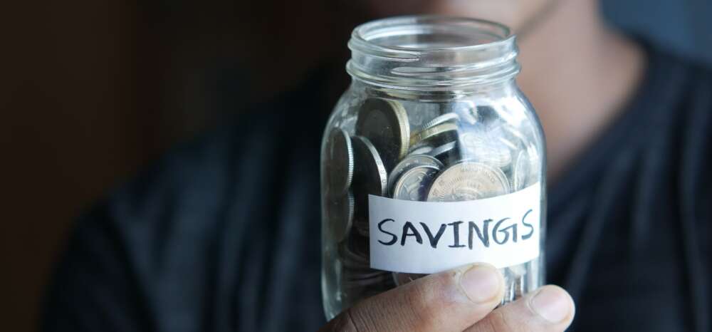 a hand holds a glass jar filled with coins labelled 'Savings'