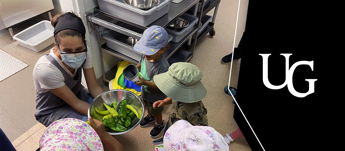 a person wearing an apron and mask bends down with a bowl of peppers as children in sun hats gather around her